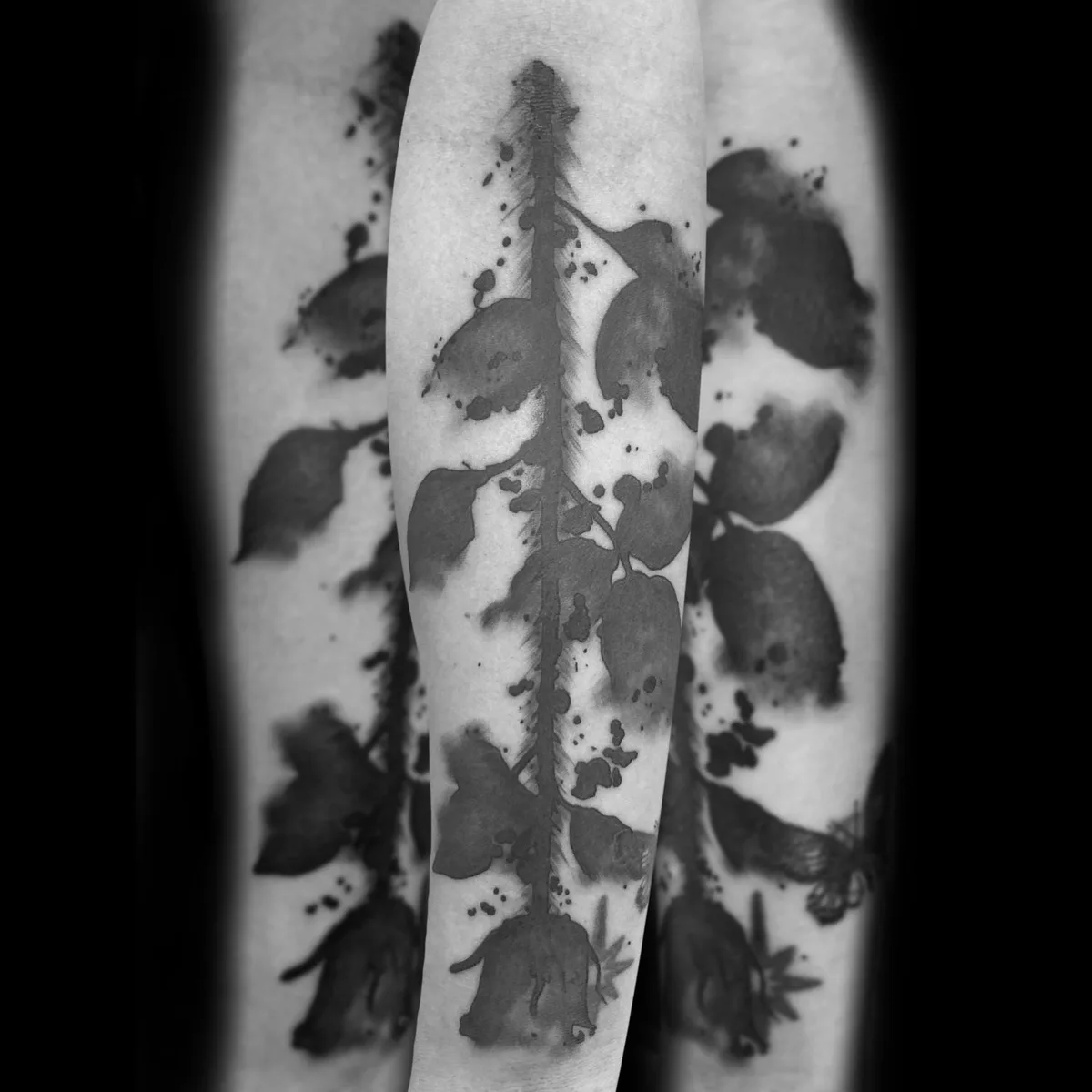 A monochromatic black and grey tattoo on an arm depicting a stylised rose stem with abstract silhouette-like blossoms and leaves, surrounded by ink-splash and splatter effects that give the edges a soft, watercolour-like quality.