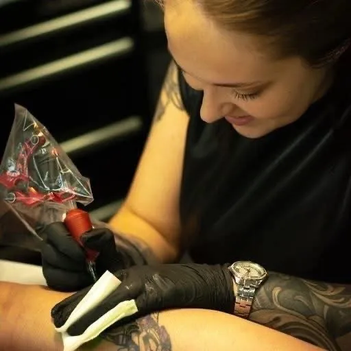 A female tattoo artist with brown hair pulled back, wearing a black sleeveless shirt and black nitrile gloves, focuses on tattooing a client's forearm. She holds the tattoo machine in her right hand and a white paper towel in her left. The client's arm and shoulder are already covered in large black