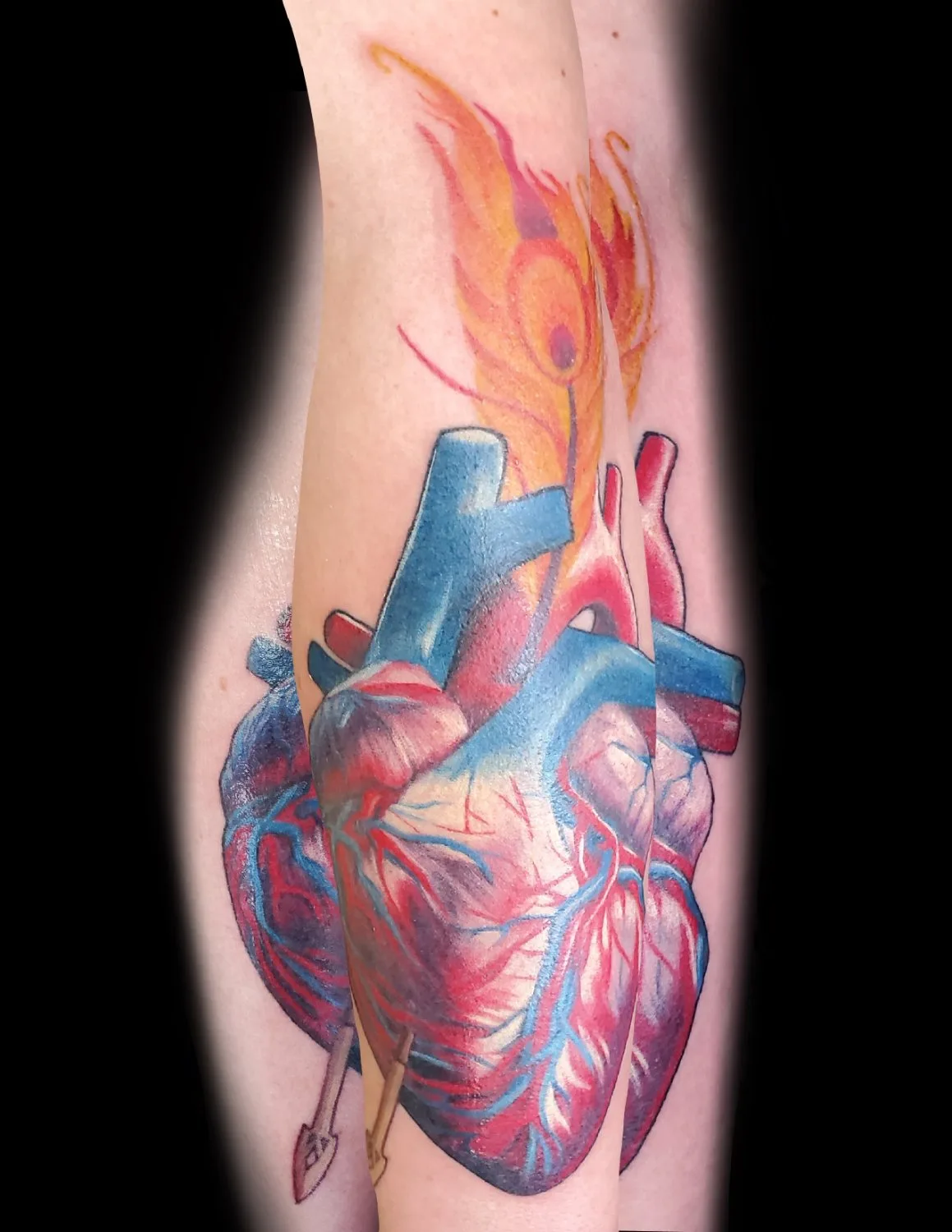 A close-up of a realistic anatomical heart tattoo rendered in deep reds and vibrant blues, with a flame-like element rising from its top and a metallic gold arrow piercing its lower portion, set against a black background.