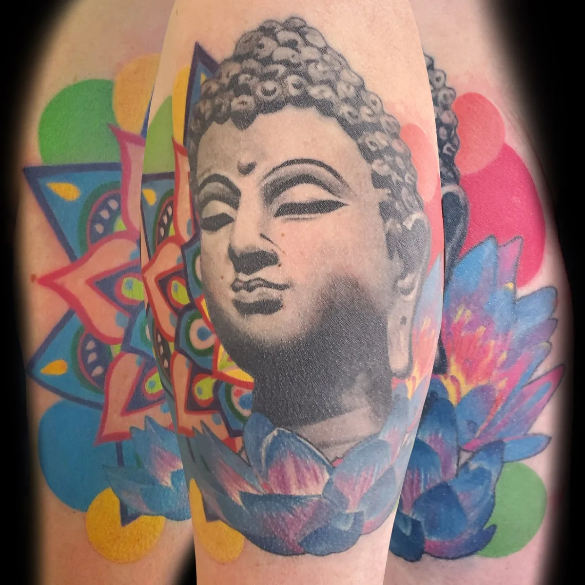A tattoo on an arm or shoulder featuring a grayscale realistic portrait of the Buddha's face with closed eyes, surrounded by a vibrant abstract design incorporating colourful lotus-like forms in bright blues, pinks, greens, and yellows.