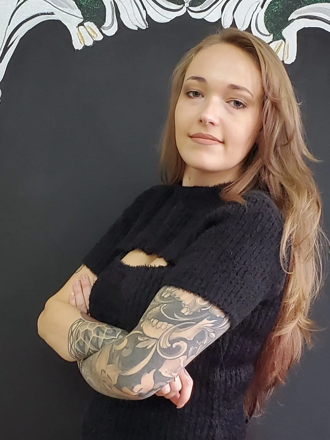 A young woman with long wavy brown hair stands with arms crossed, looking directly at the camera. Her left arm is heavily tattooed with intricate black ink designs. The background is dark charcoal grey with decorative floral artwork in the corners.