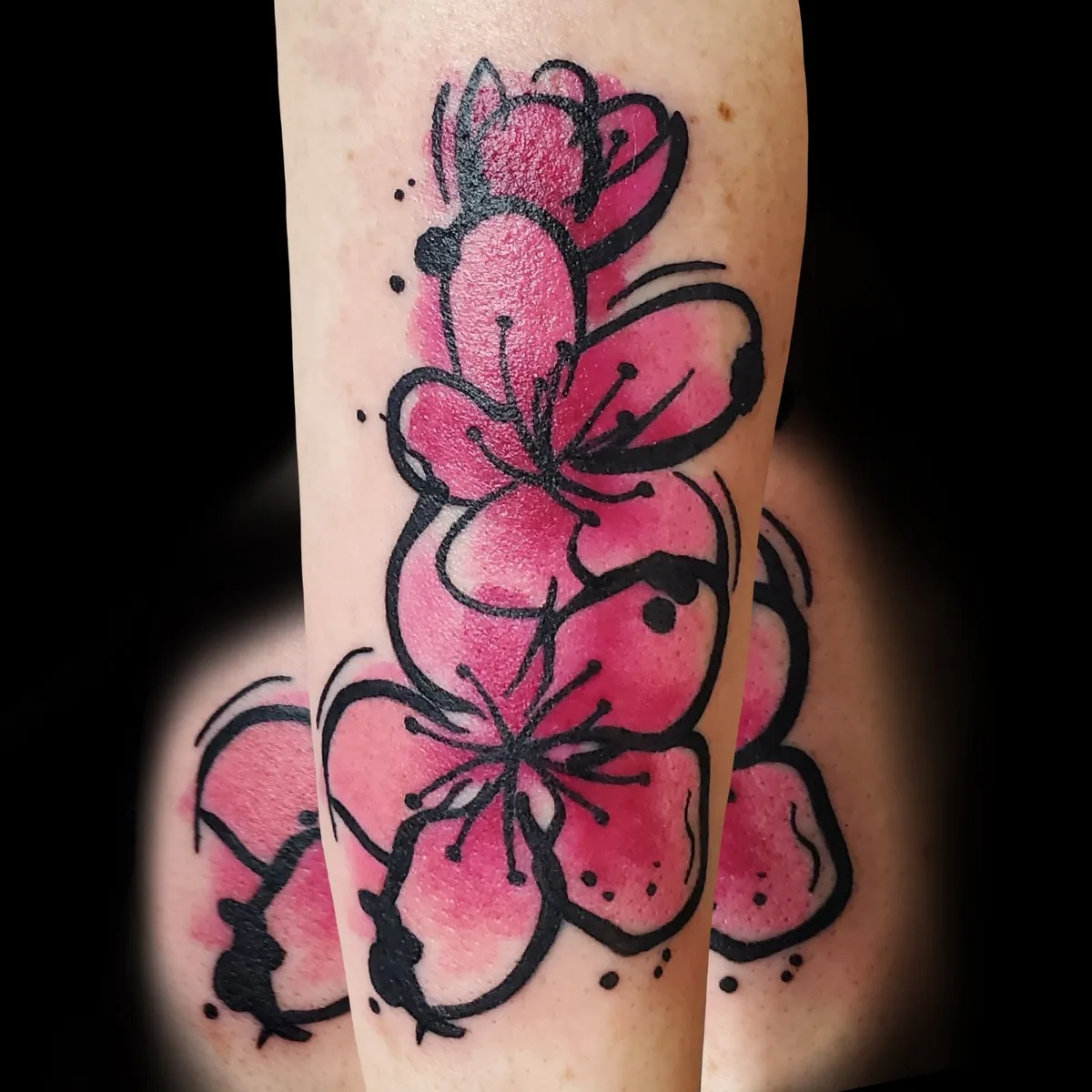 A tattoo of cherry blossoms on an arm, rendered in bold black linework with vibrant pink watercolour washes and scattered black ink splatters, creating a cascading floral composition against a dark background.