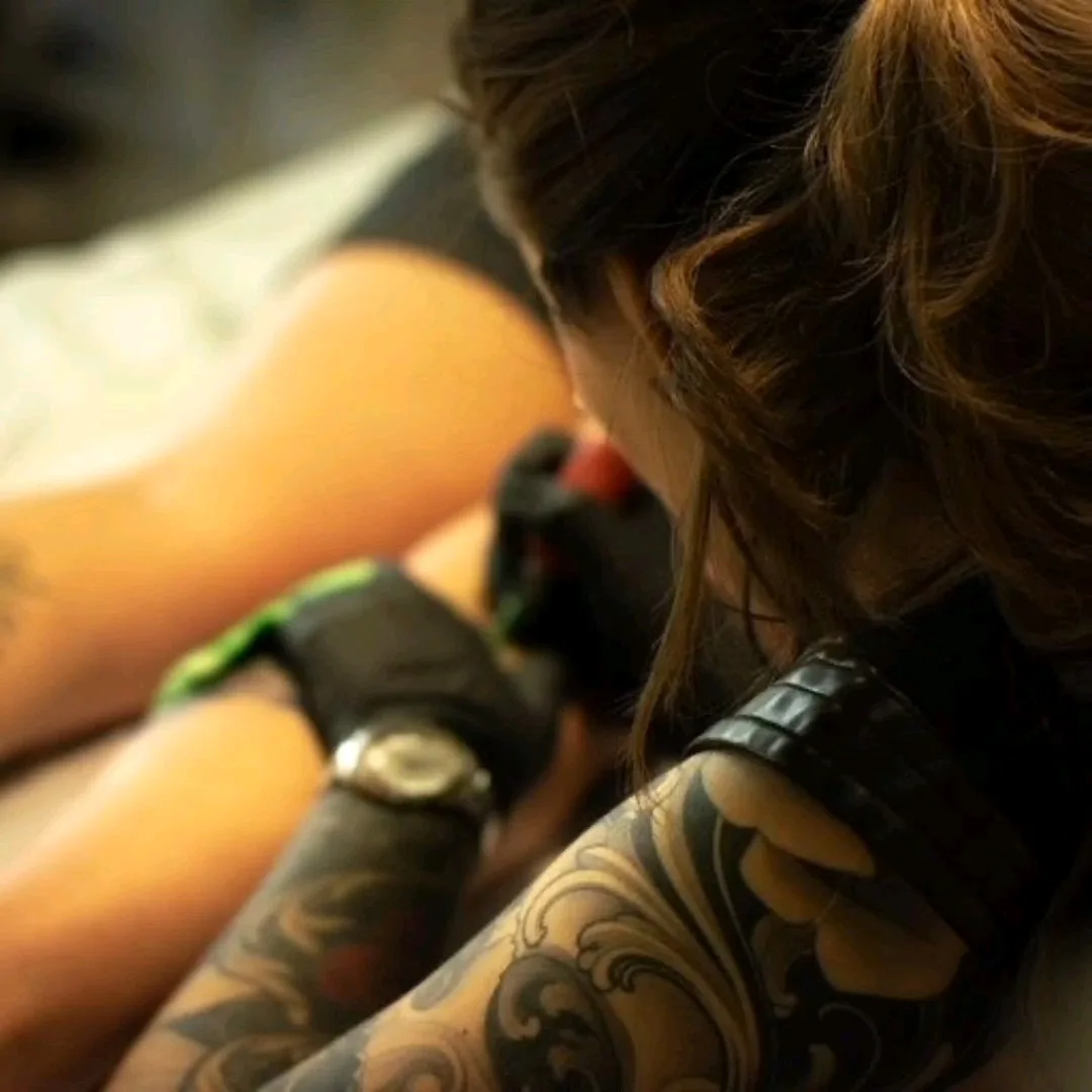 A candid close-up of a tattoo artist in profile, looking down as she inks a client's leg. The artist wears black gloves and has a heavily tattooed arm visible in the foreground. The client's leg bears large swirling wave-like tattoo motifs in black ink. Warm artificial light illuminates the working 