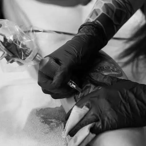 A black and white close-up of a tattoo artist's gloved hands. The right hand holds a tattoo machine with its needle close to the client's skin, which already bears a circular tattooed motif. The left hand holds a white paper towel. The artist's own arm displays a geometric tattoo pattern.