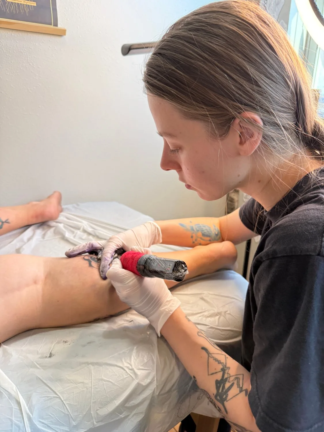A tattoo artist with light brown hair in a ponytail bends over a client's leg, holding a tattoo machine in white latex gloves. The client lies on their side on a white table. The artist wears a black t-shirt. A framed artwork is partially visible on the wall behind.