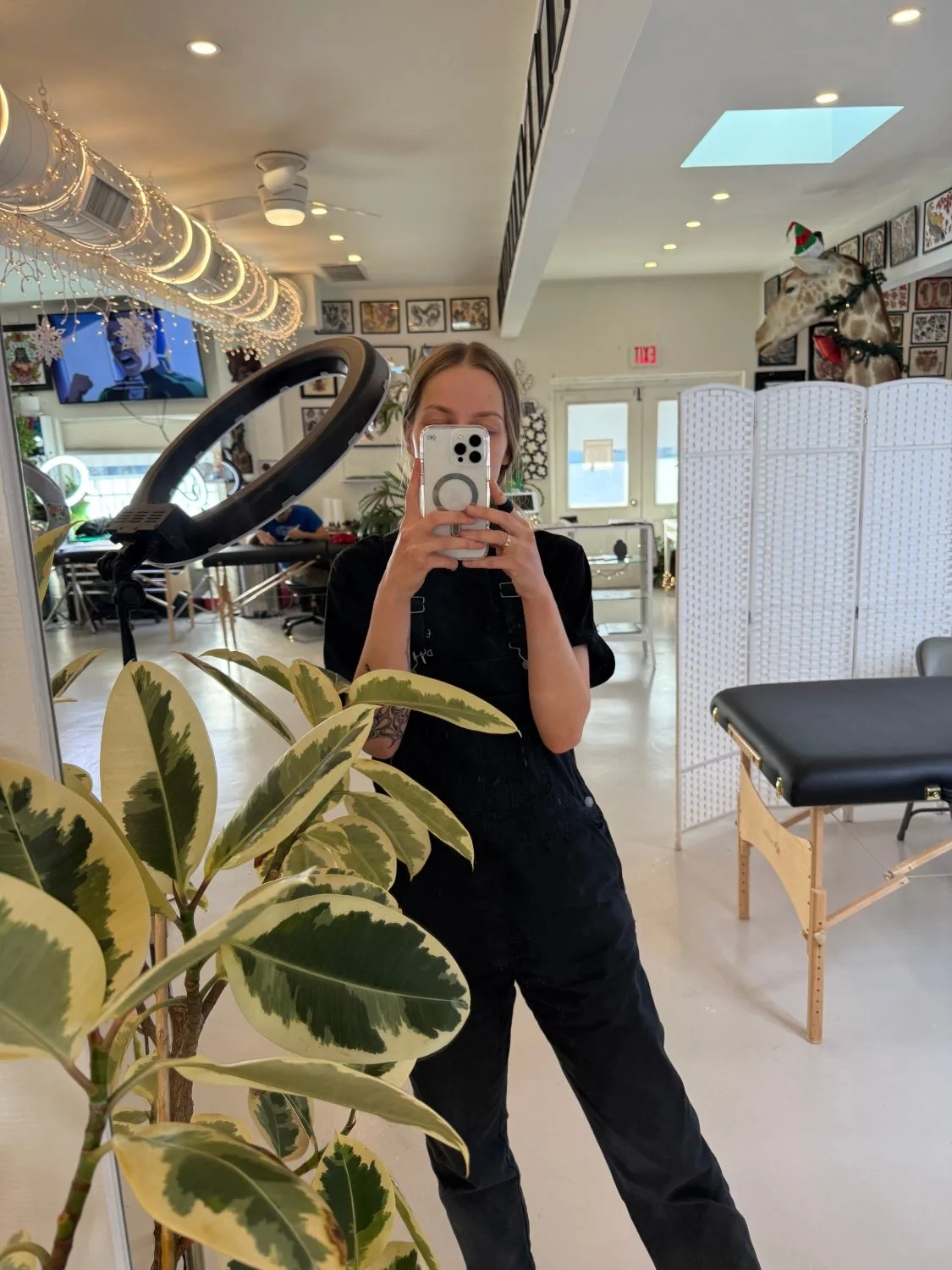 A mirror selfie in a bright studio. A woman in black overalls holds a smartphone at chest height. Behind her reflection: framed tattoo designs on white walls, a large ring light, a massage table, plants in the foreground, string lights, and a taxidermied giraffe head wearing a small hat and garland