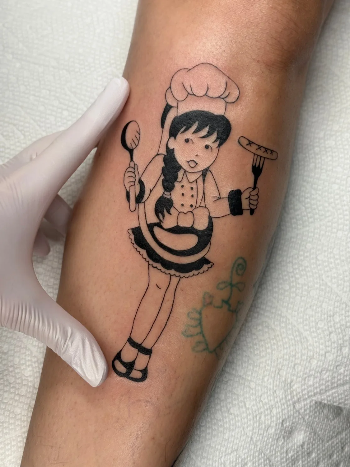 A black ink tattoo on skin showing a cartoon girl dressed as a chef. She wears a tall puffy hat, a dotted white jacket with dark collar and cuffs, a black frilled apron, and holds a fork with a sausage and a large spoon. Her hair is in a braid. A gloved hand is visible at the upper left edge of the