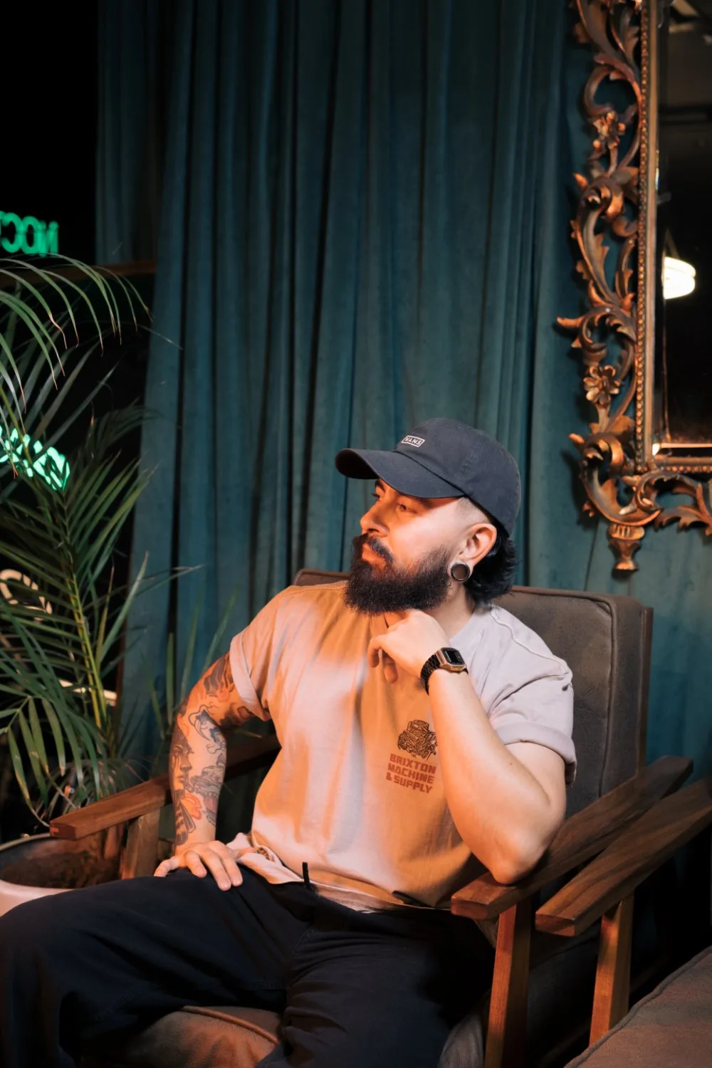 A man with a full beard, dark mullet hair, and a black baseball cap sits in a dark brown upholstered armchair. He wears a light tan Brixton t-shirt and dark pants. His left arm rests upward with his hand near his chin, showing colorful traditional-style tattoos on the bicep and forearm. A large pott