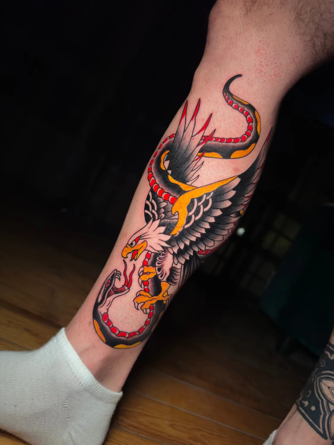 A traditional-style tattoo on a lower leg depicting an eagle with white head, black wings tipped in red, and yellow talons gripping a black-and-yellow snake that coils around its body. Bold black outlines and a flat, graphic color palette define the composition.