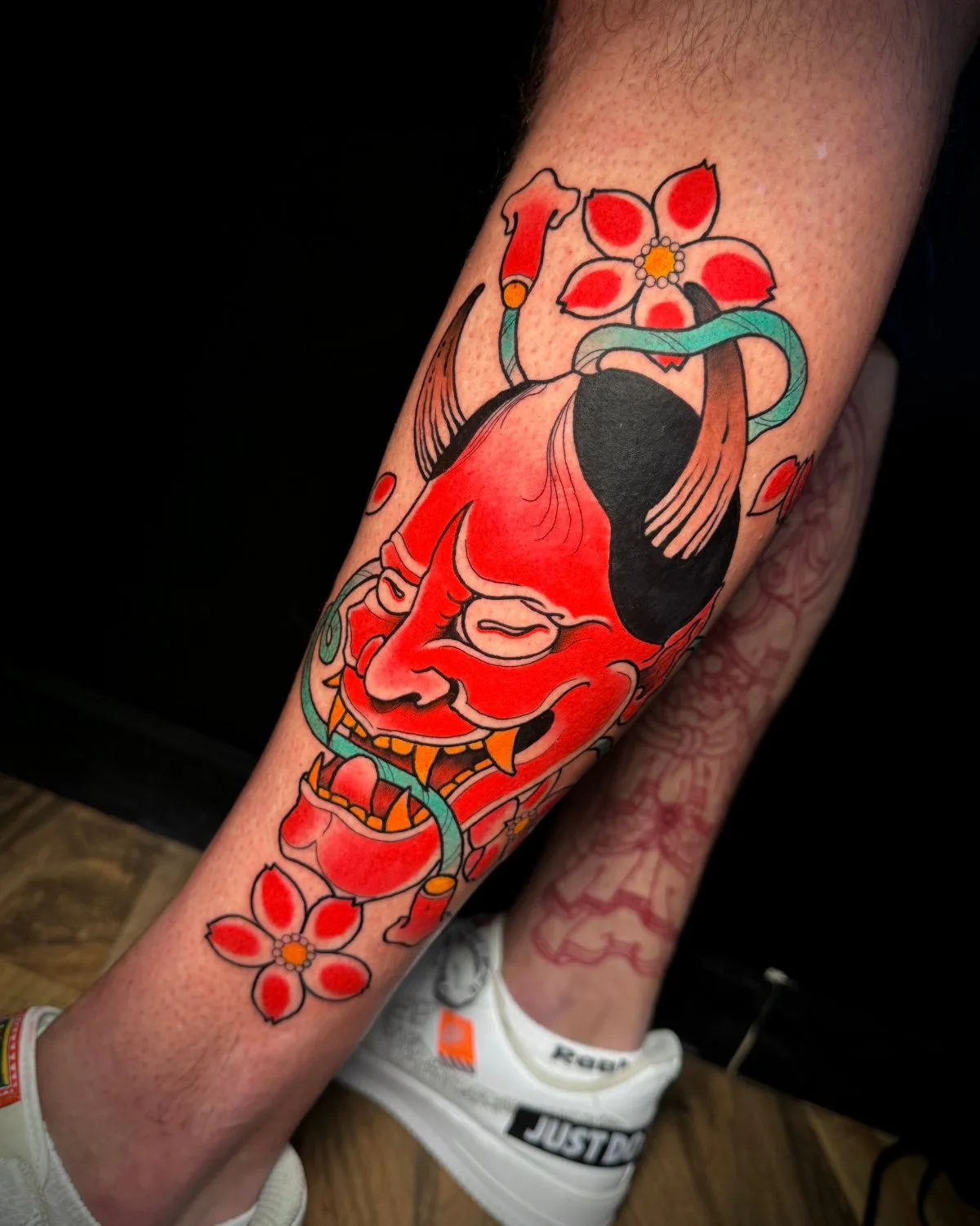 A tattoo on a lower leg showing a red Oni mask with a teal snake coiled around it and red cherry blossom flowers, executed in a bold neo-traditional Japanese style with solid colour fills and thick black outlines.