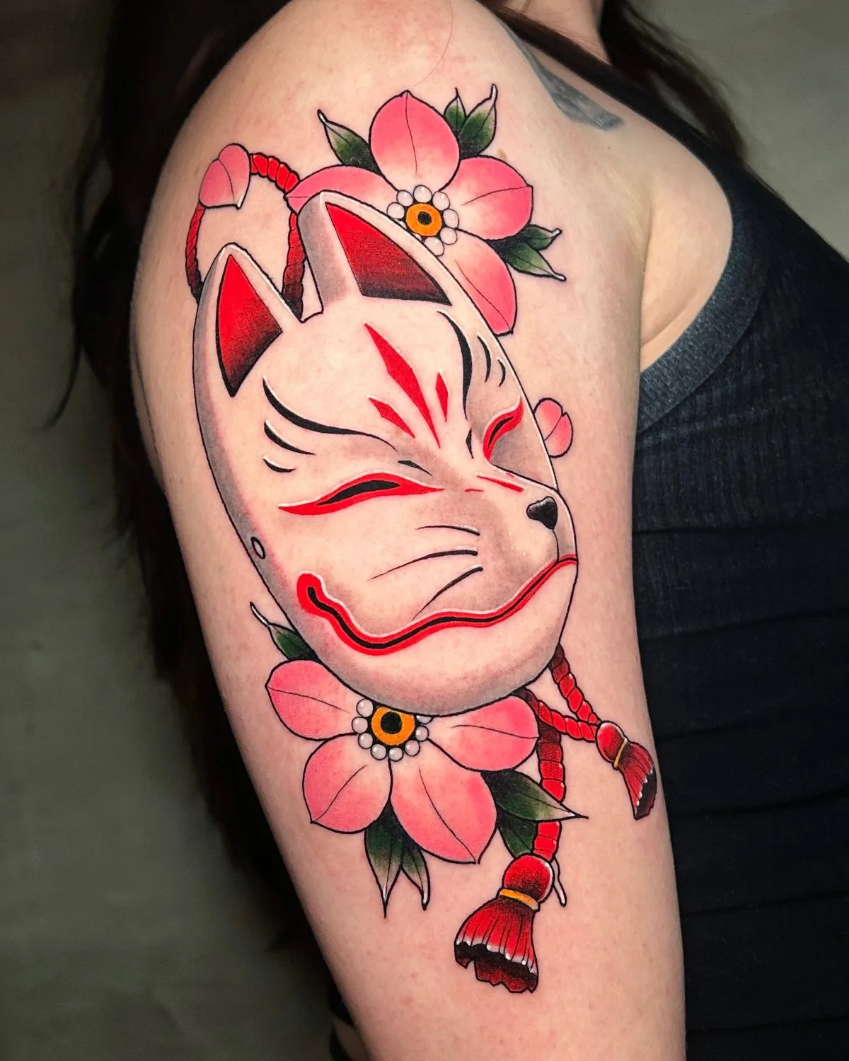 A tattoo on the upper arm showing a stylized white Kitsune fox mask with red eye markings, black whiskers, and pointed ears. Two soft pink cherry blossoms with yellow-orange centers are placed above and below the mask. A vivid red braided cord with hanging tassels connects the elements. Bold outline