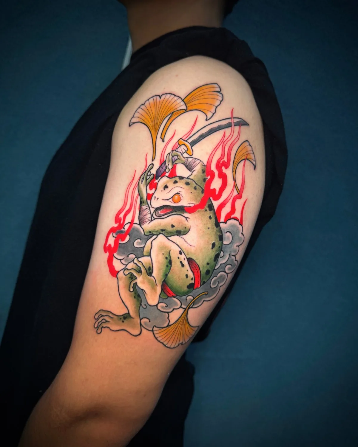 A tattoo on the upper arm showing a green frog in a dynamic upright pose, holding a katana with a gold hilt raised above its head. Red flames swirl around it, soft grey clouds anchor the lower composition, and fan-shaped golden ginkgo leaves frame the upper portion. The frog's limbs are wrapped in p