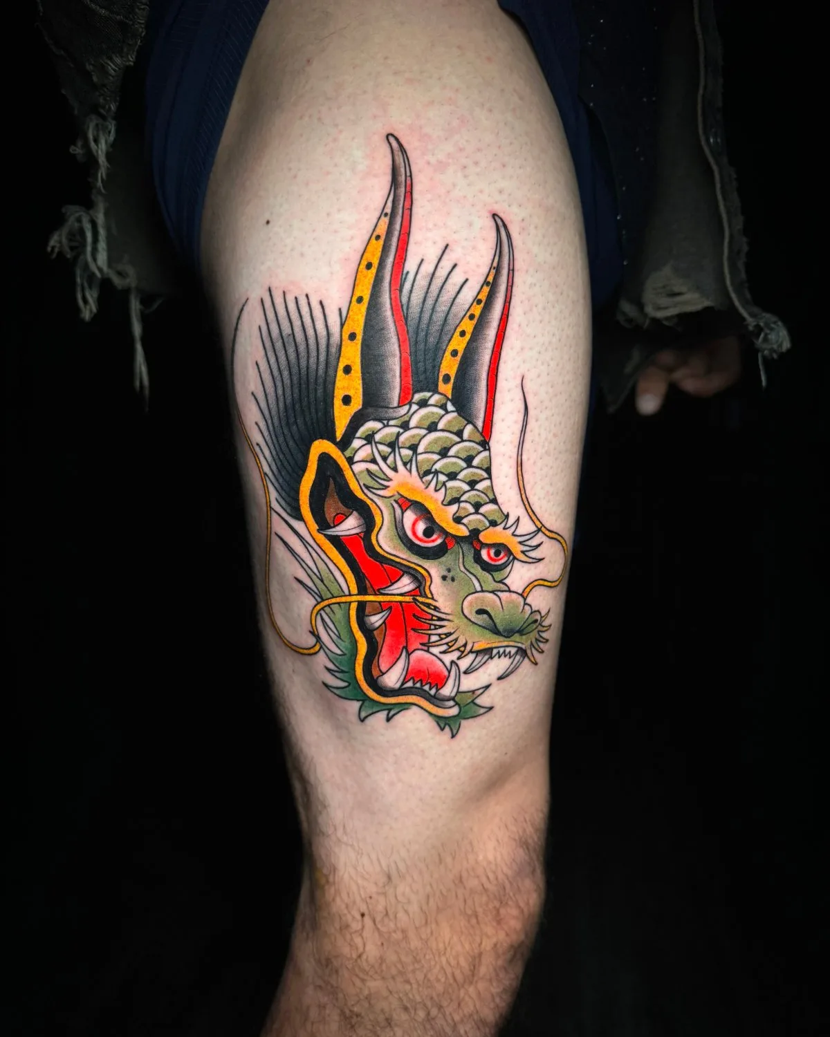 A tattoo on the thigh depicting the head of a dragon in traditional American style. The face is green with lighter scale accents, the mouth is open showing white teeth and a red-lined interior, and two horns are tipped in yellow. A spiky black mane extends upward and to the left. Bold black outlines