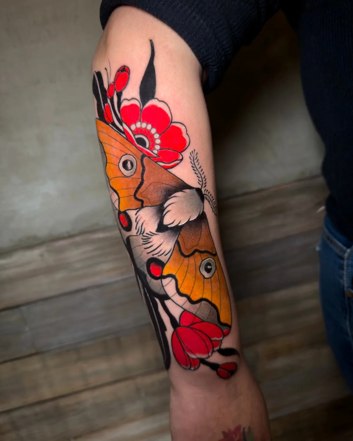 A tattoo on an arm depicting a moth with wide-spread wings that shift from rich orange at the top to lighter yellow-orange at the edges, with brown-grey patterning and a circular eye-spot on each upper wing. Several vivid red flowers with black outlines and white-accented centers are arranged around