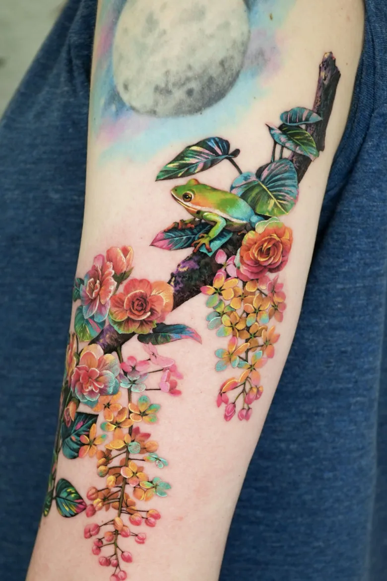 Frog on a Branch Tattoo – A tattoo of a bright green frog sitting on a branch with flowers. The composition includes intricately detailed leaves and a realistic depiction of a nighttime sky.