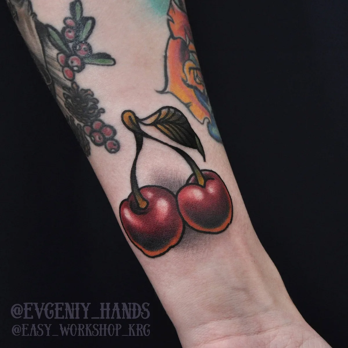 A traditional tattoo of two cherries joined at the stem, rendered with realistic shading and vivid red and green colour. The design is bold and classic.