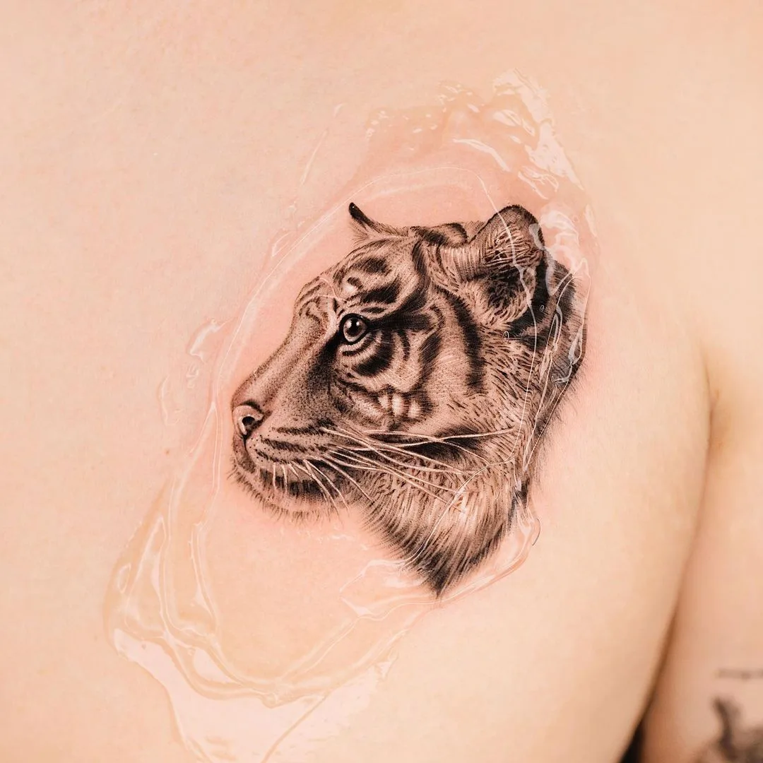 A detailed black and grey tattoo of a tiger's head in profile. The tattoo appears fresh, with a clear protective layer over it.