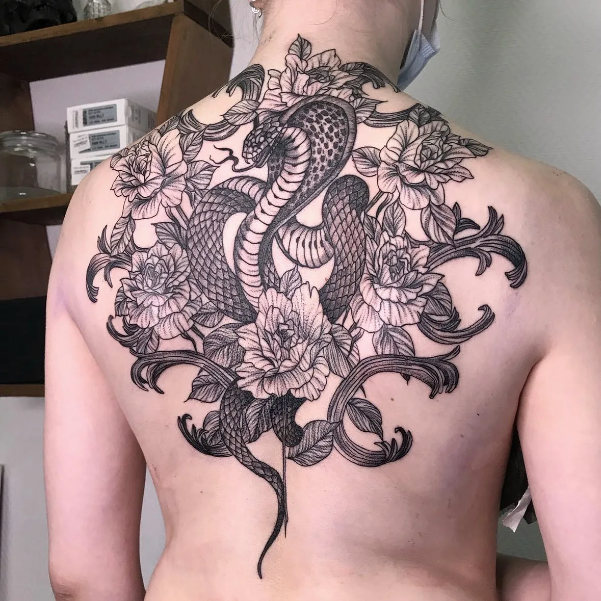 A detailed black and grey tattoo of a cobra with its hood partially spread, intertwined with flowers and ornamental linework.