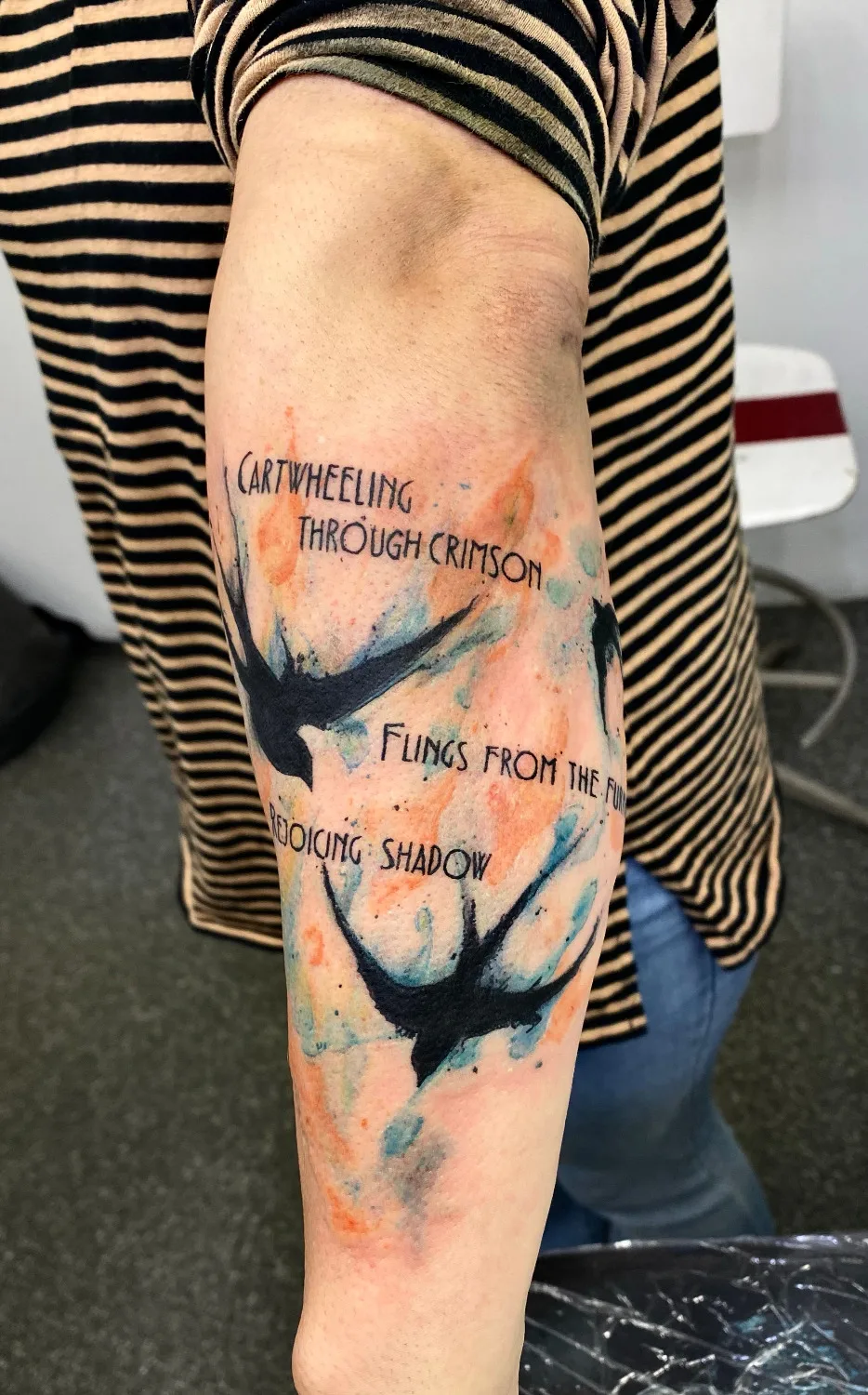 A watercolour-style tattoo featuring swallows with colour bleeding past loose outlines, accompanied by lines of poetic text.