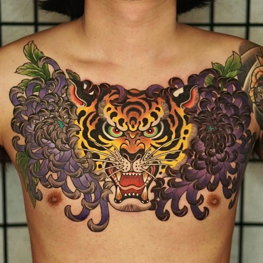 A vibrant Japanese-style tattoo of a detailed tiger head surrounded by large chrysanthemum flowers in rich oranges and yellows.