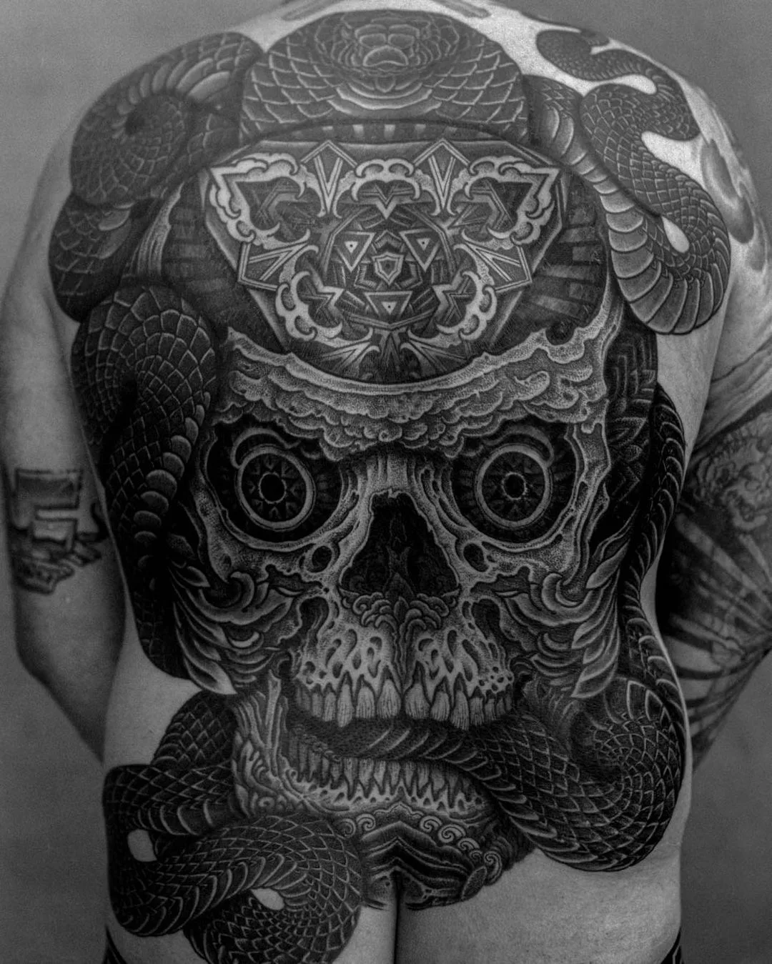 A large, intricate blackwork tattoo featuring a skull intertwined with snakes and geometric patterns.
