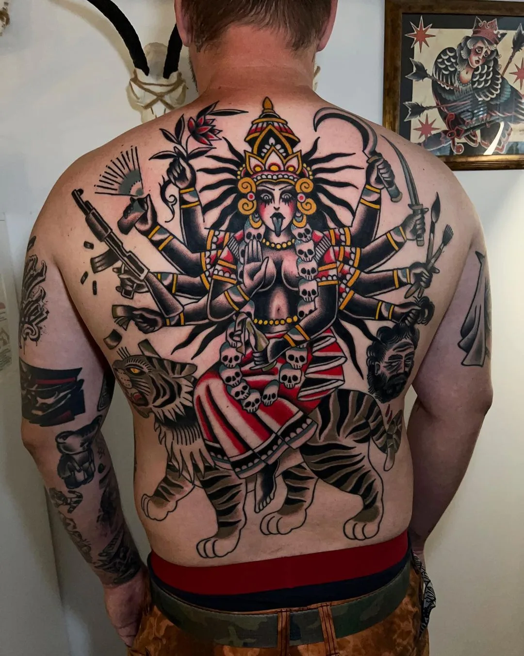 A large, colorful traditional tattoo of a multi-armed deity riding a tiger, holding various weapons, with skulls incorporated into the design.