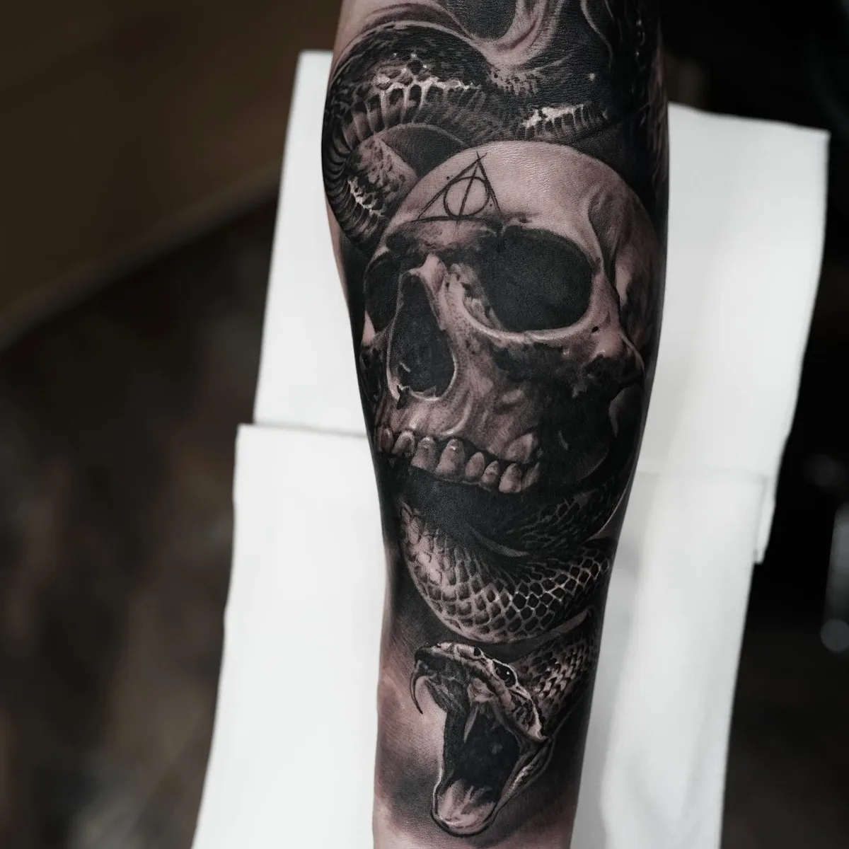 A detailed black and grey tattoo featuring a realistic skull intertwined with a snake, with fine shading and texture throughout.