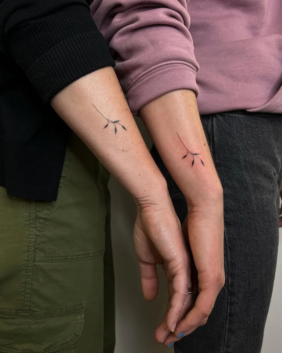Two forearms each bearing a small, minimalist tattoo of a simple plant sprig with fine black linework. The designs are matching and placed symmetrically.