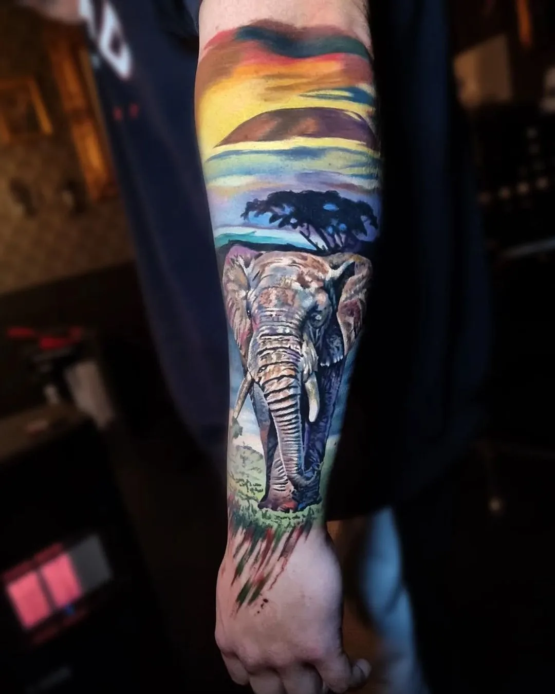 A vibrant full sleeve tattoo depicting a realistic elephant in a savanna landscape beneath a warm, colourful sunset sky with rich oranges and yellows.