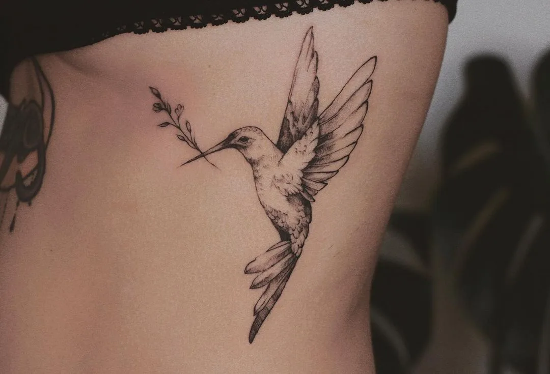 A small, delicate black and grey fine-line tattoo of a hummingbird carrying a sprig in its beak, placed on the ribcage.