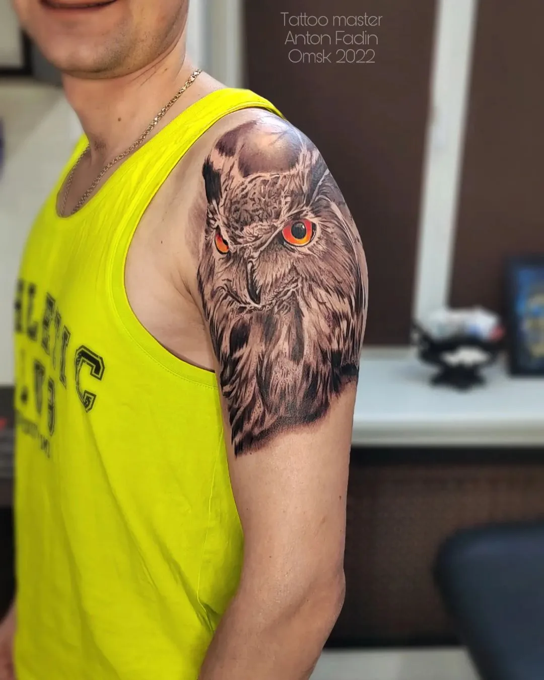 A black and grey realistic owl portrait tattoo on the upper arm, featuring vibrant orange eyes and detailed feather shading.