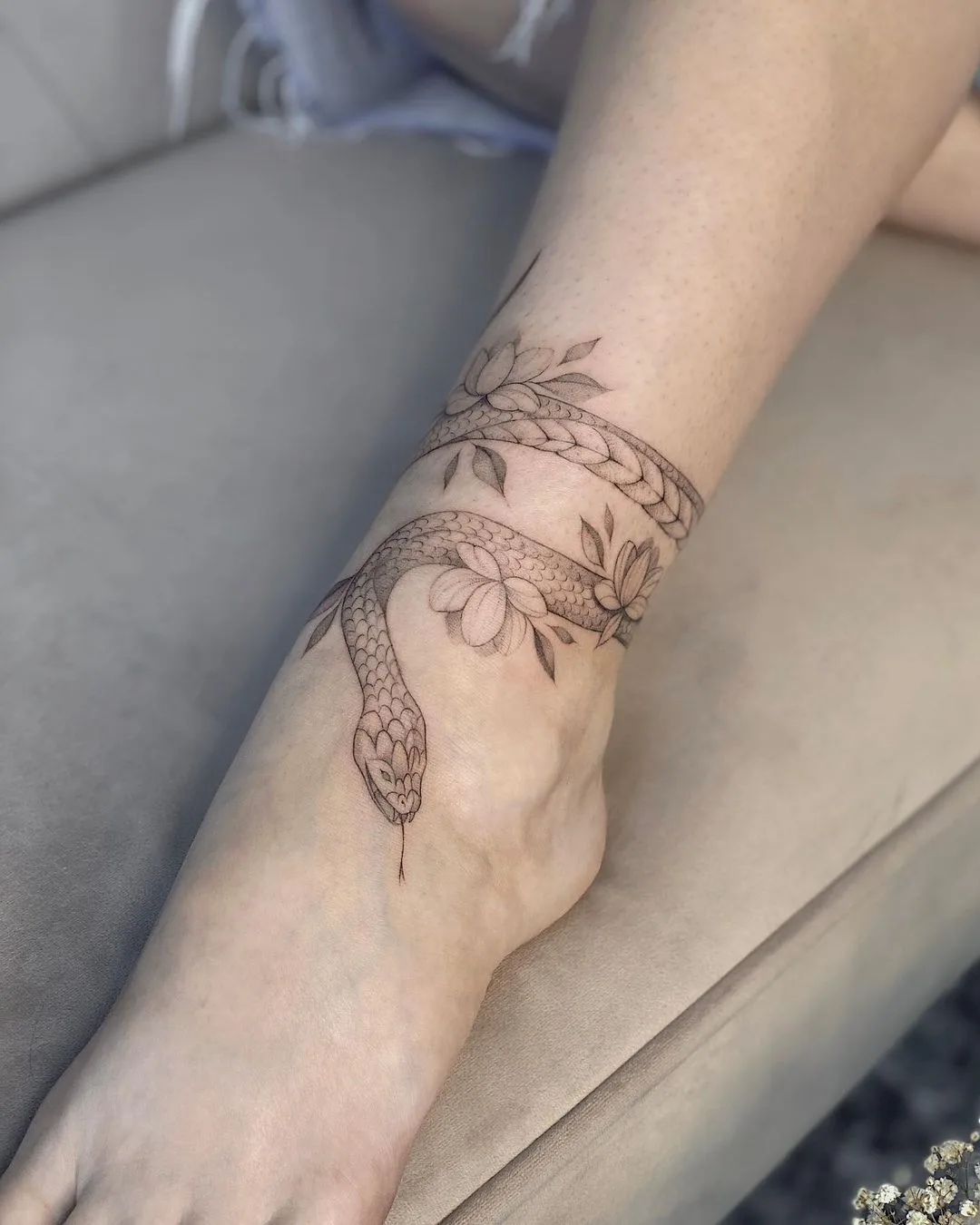 A delicate black and grey fine-line tattoo of a snake wrapped around the ankle, accompanied by small floral elements.