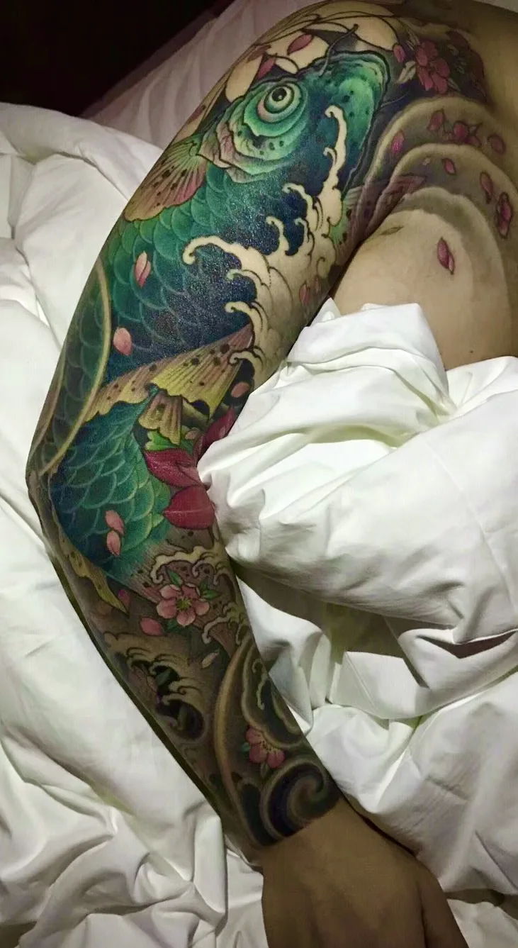 A full sleeve tattoo in Japanese style showing a koi fish swimming through waves with cherry blossom petals scattered throughout.