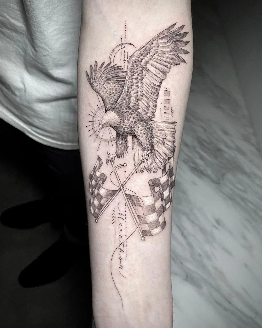 Black and grey forearm tattoo of an eagle holding checkered flags with geometric shapes and script lettering.