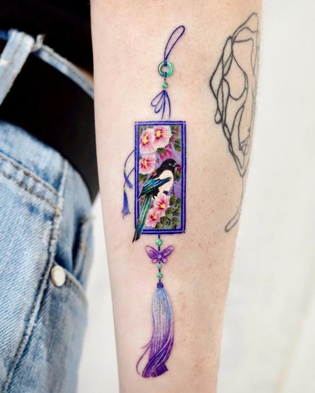 A colorful tattoo featuring a bird, flowers, and a tassel design in a Japanese-inspired style.
