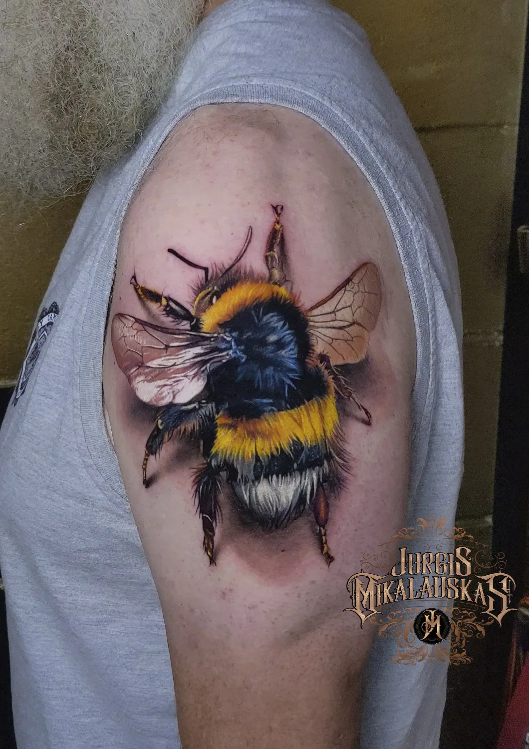 A hyperrealistic tattoo of a bumblebee on the shoulder, with highly detailed fur texture, translucent wing veins, and precise shading that makes the insect appear three-dimensional against the skin.