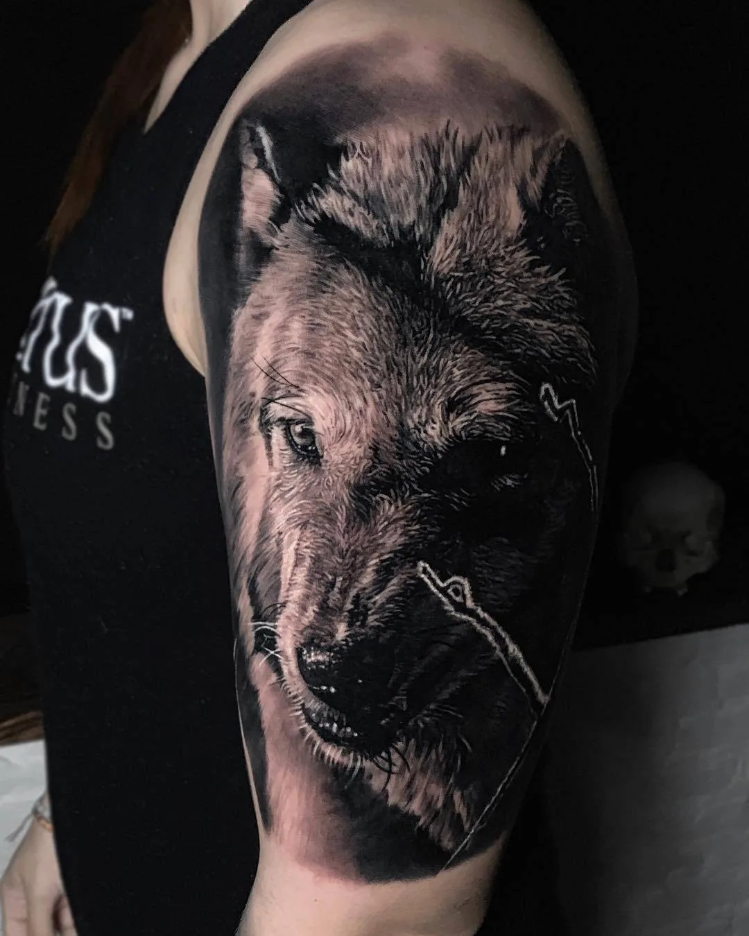 A detailed black and grey tattoo of a wolf's face on the upper arm. The tattoo showcases impressive realism and shading techniques.