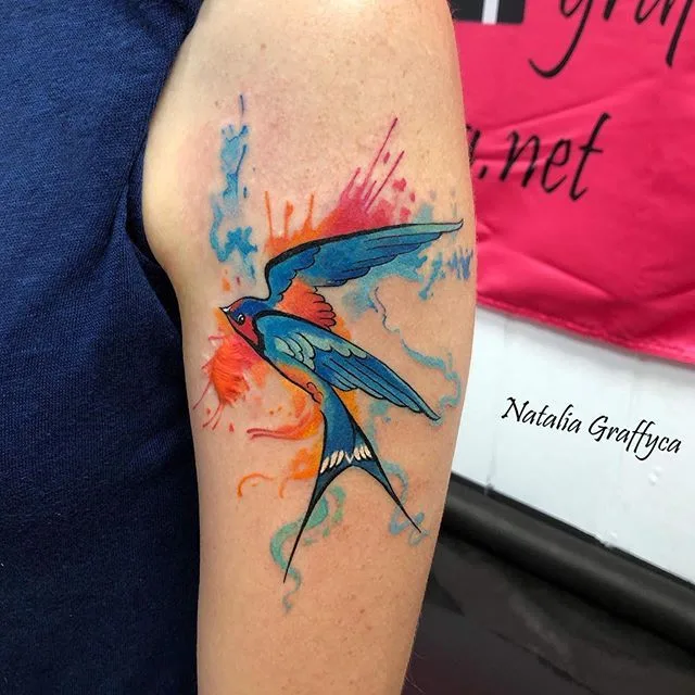 Tatouage aquarelle vibrant d'une hirondelle en vol entourée d'éclaboussures de peinture colorées.