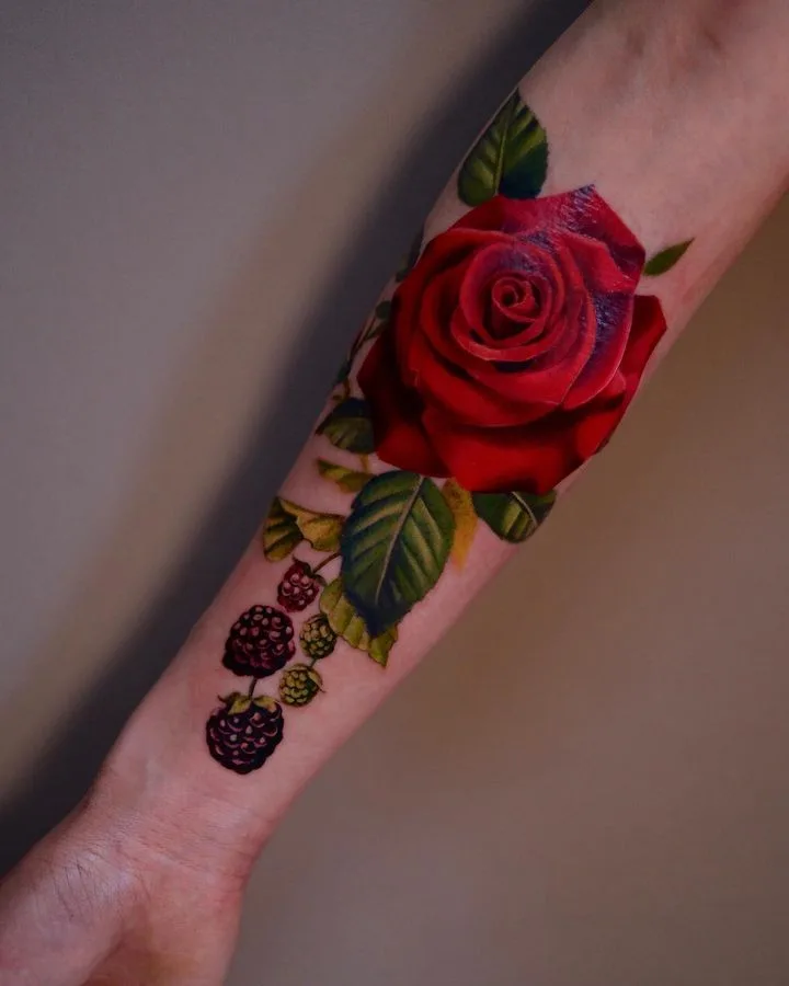 Ein detailliertes Tattoo einer roten Rose und Brombeere mit grünen Blättern, in lebhafter, realistischer Farbe auf der Haut dargestellt.