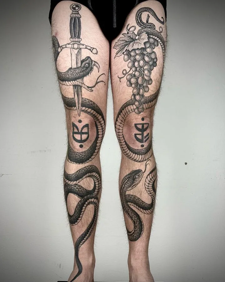 A detailed black and grey tattoo on the knees featuring two snakes intertwined around a sword with grape clusters, referencing caduceus and rod of Asclepius imagery.