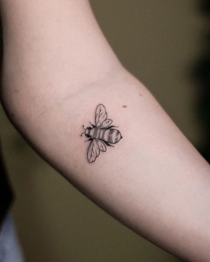 A small, precisely rendered bee tattoo in black ink using a linework style, showing clean outlines of the body, wings, and legs with minimal fill.