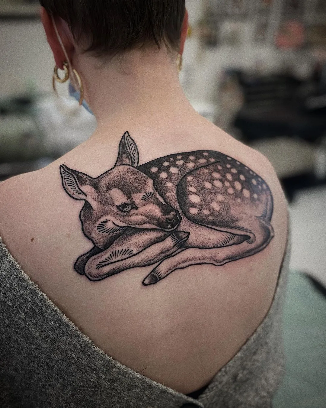 A detailed black and grey tattoo of a resting fawn on the shoulder blade, rendered with realistic shading and dotwork technique.