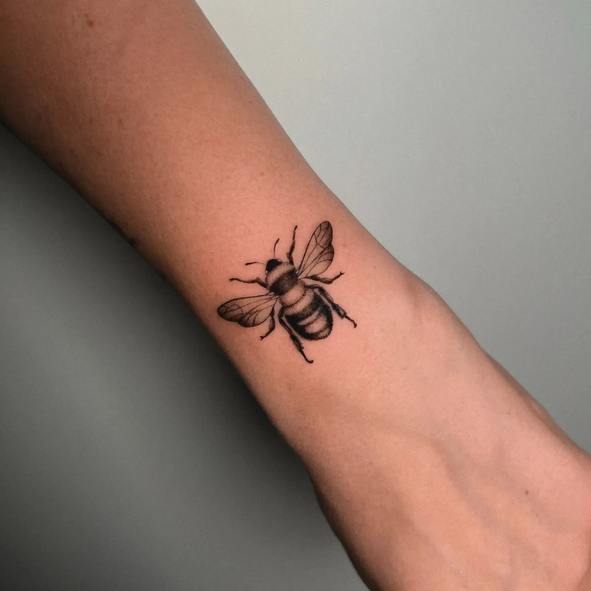 A detailed black and grey bee tattoo on the forearm rendered in a fineline style, with delicate outlines and minimal shading showing the insect's wings, body, and antennae.