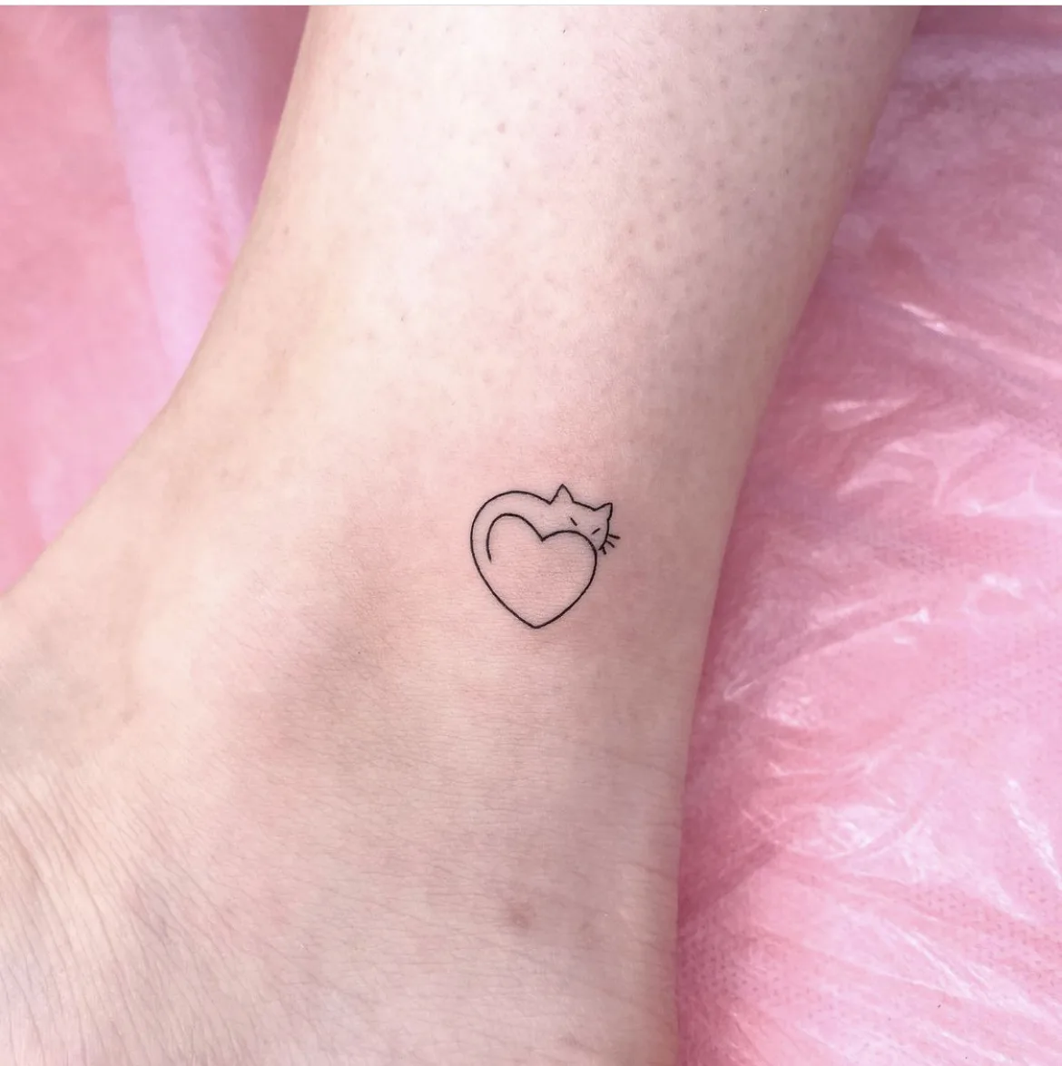 A simple and elegant minimalist black tattoo of a cat curled into a heart shape, rendered in clean linework on pale skin.