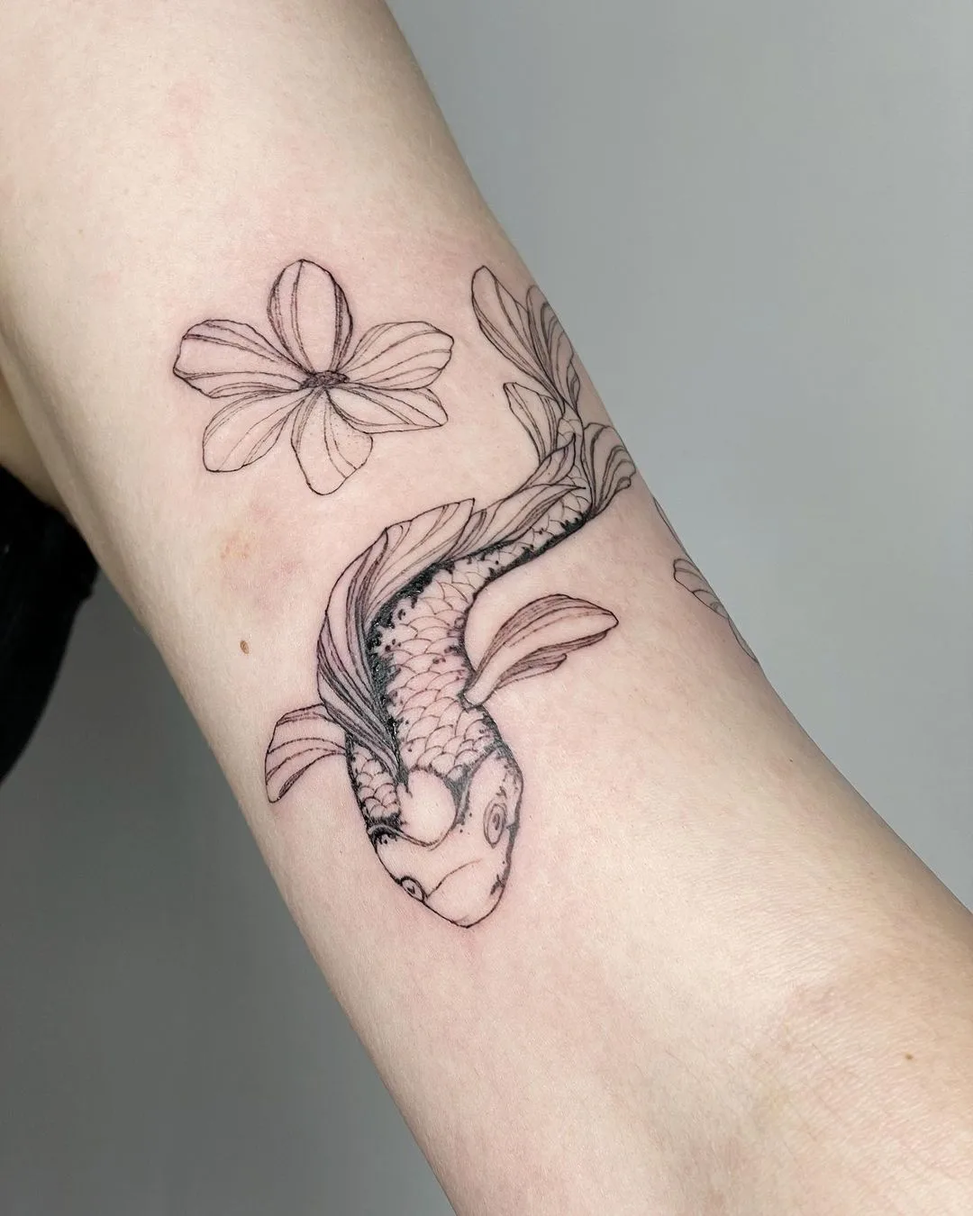 A delicate black ink linework tattoo on skin showing a koi fish alongside a small flower in a minimal style.