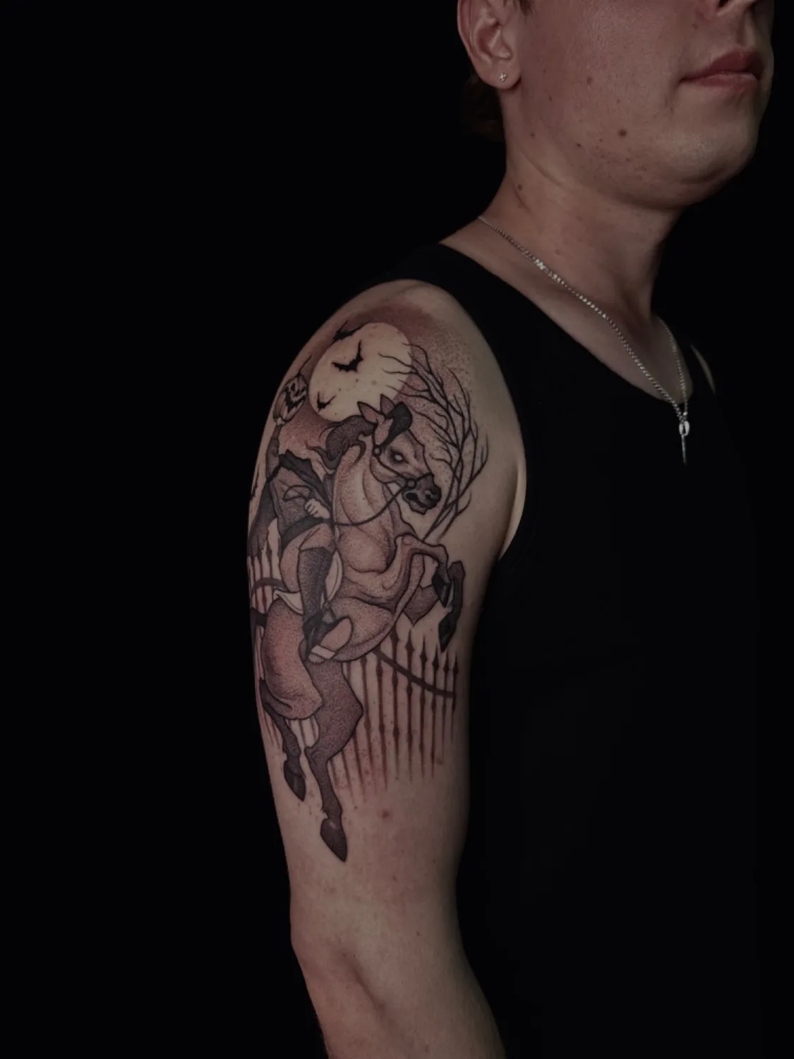 A black and grey tattoo depicting the Headless Horseman riding a horse in front of a fence and a full moon.