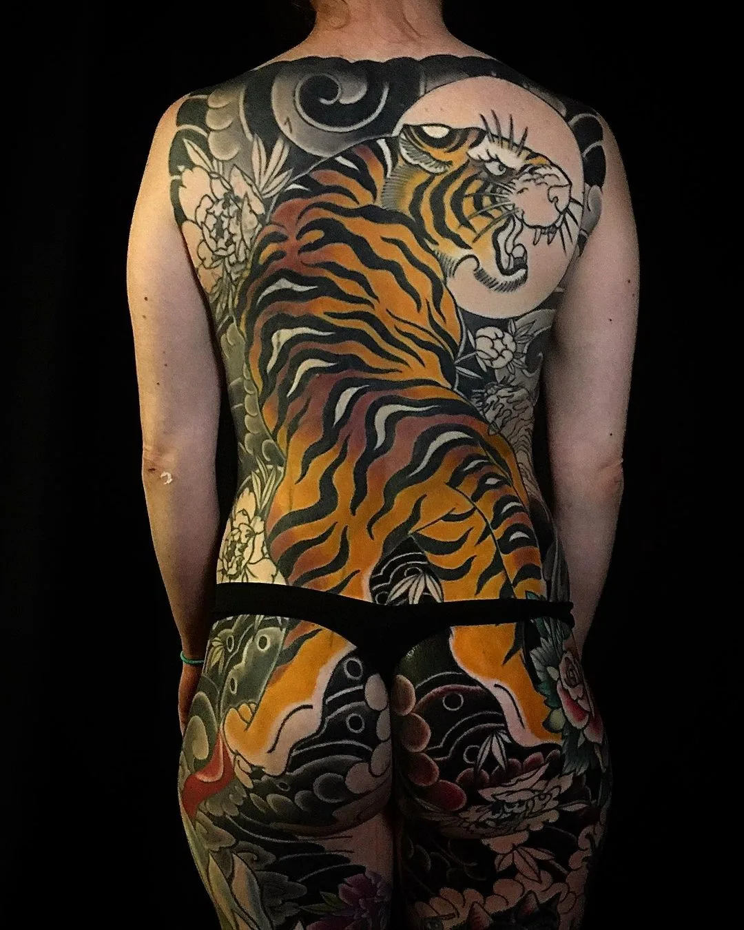 A large, colorful Japanese-style tattoo covering the entire back, featuring a tiger surrounded by flowers and clouds in vibrant reds, oranges, and greens.