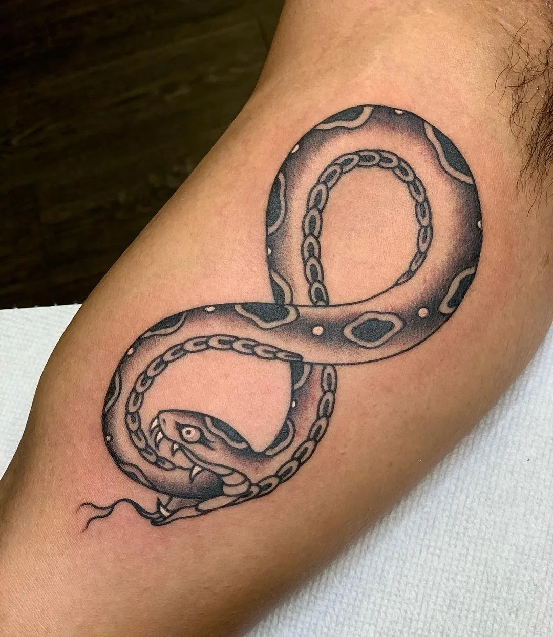 A traditional-style black and grey tattoo of a snake arranged in an infinity symbol shape, referencing the ouroboros cycle of renewal.