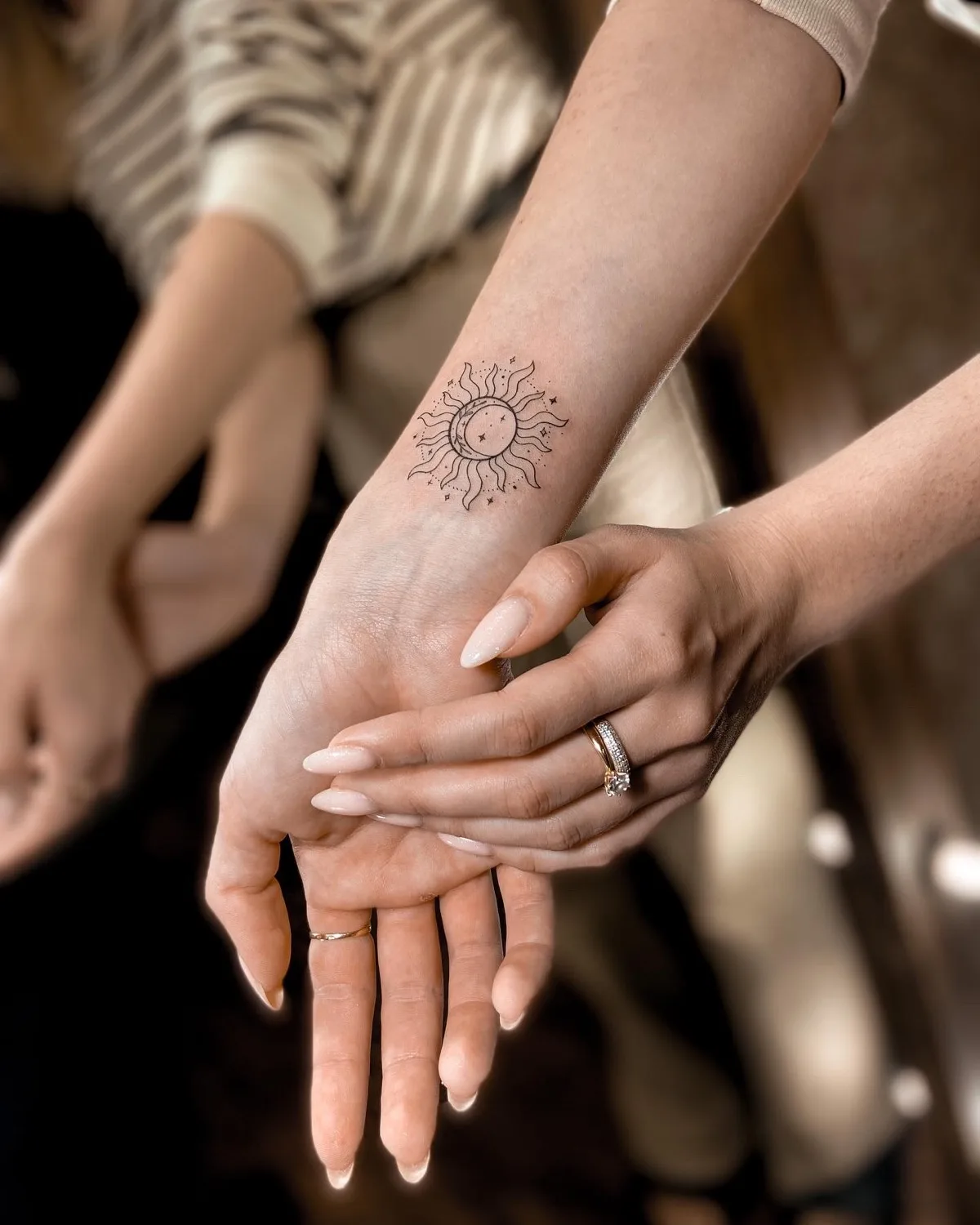A single forearm bearing a delicate linework tattoo combining a sun and a crescent moon with scattered stars. Fine black lines on pale skin.