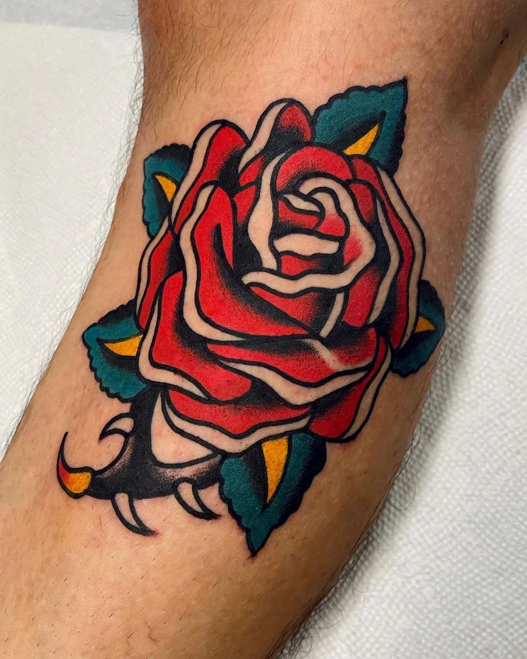 Ein klassisches Rosen-Tattoo im traditionellen Stil mit dicken schwarzen Umrissen, leuchtend roten Blütenblättern und grünen Blättern.